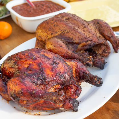 Two roasted chickens on a platter with a side of sauce and bread in the background.
