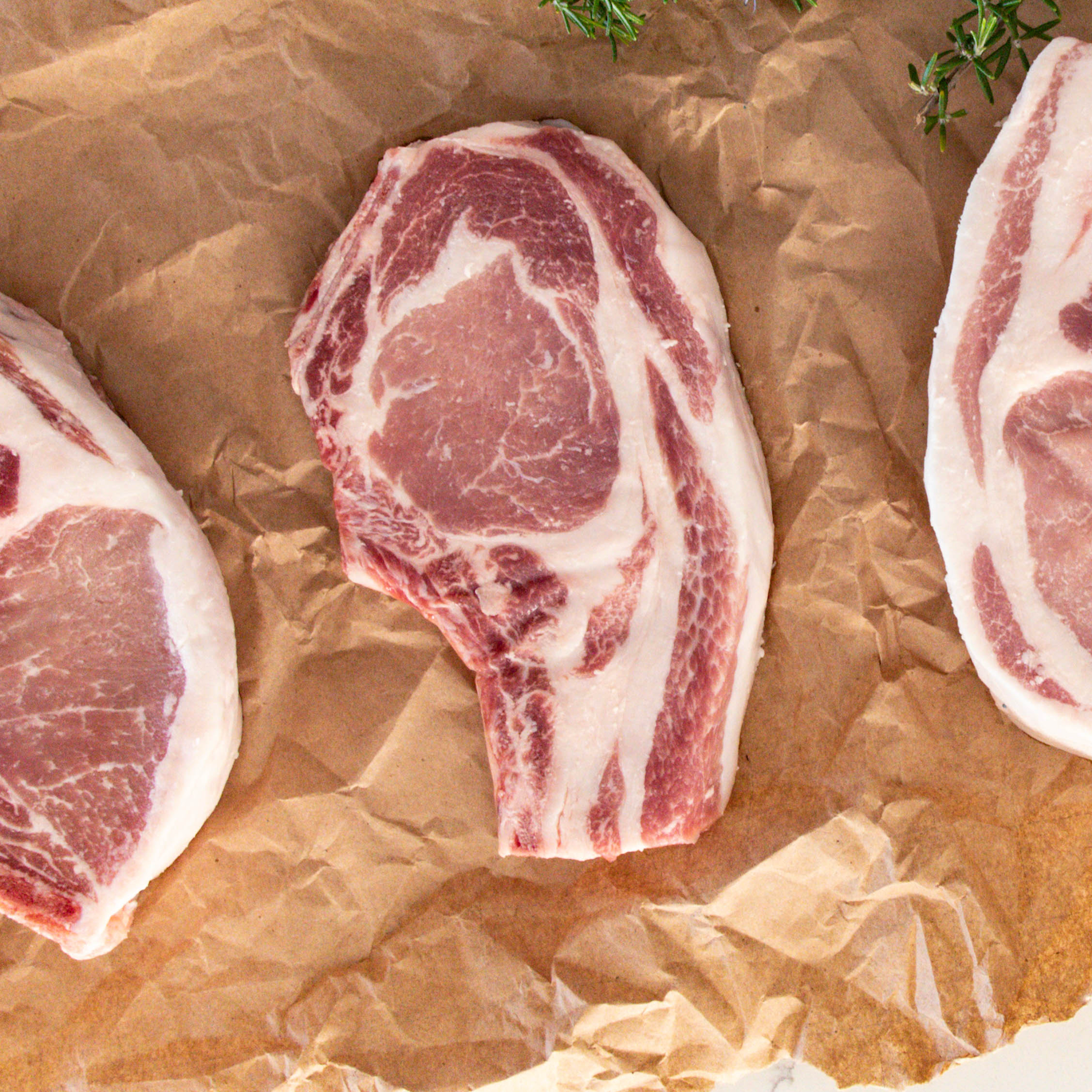Three raw pork chops on brown paper with herbs.