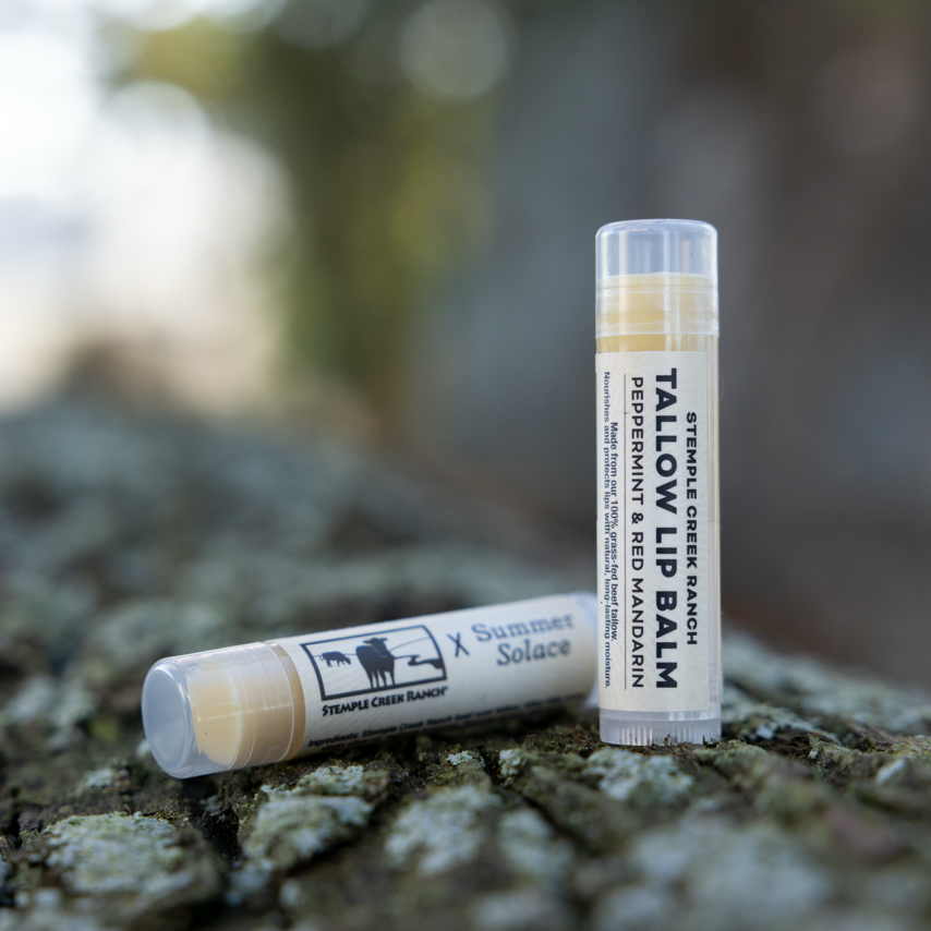 Two tubes of Tallow Balm on a textured surface with a blurred natural background